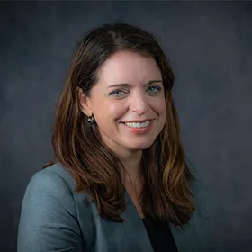 A woman with long brown hair, wearing a grey blazer and black top, smiles at the camera in front of a dark gray background.